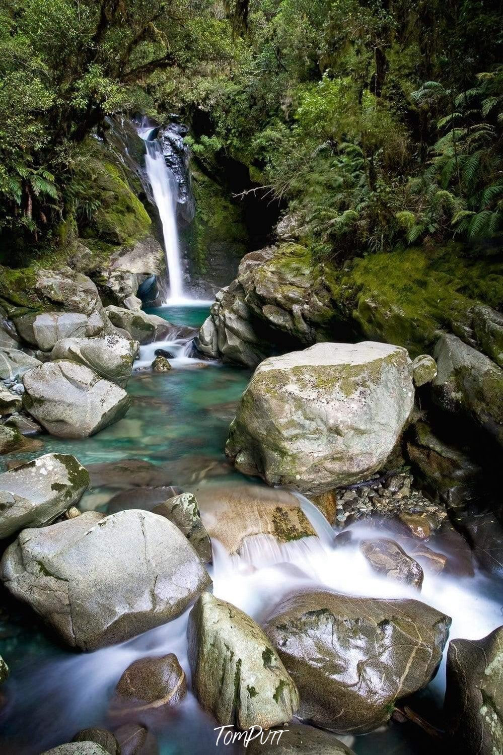 A small waterfall from a mound into a small lake surrounded by big rocky stones, Waterfall 5, Milford Track - New Zealand